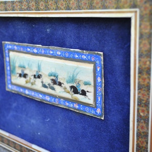 May include: A framed miniature painting depicting a scene of people riding horses in a landscape. The painting is surrounded by a decorative border with blue, white, and gold accents.