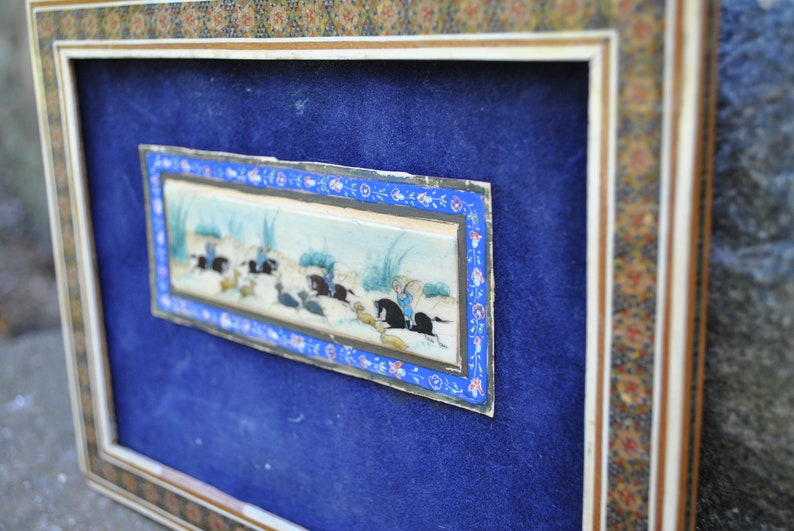 May include: A framed miniature painting depicting a scene of people riding horses in a landscape. The painting is surrounded by a decorative border with blue, white, and gold accents.