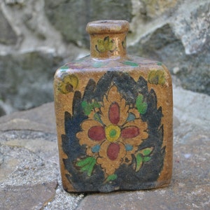 May include: A small, square, ceramic bottle with a floral design. The bottle is painted with a black, green, red, and yellow floral pattern.