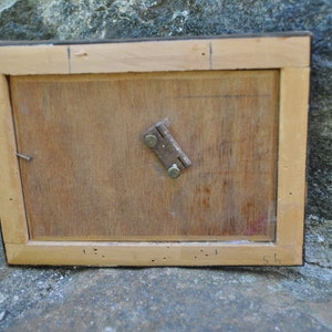 May include: The back of a wooden picture frame with a metal hinge. The frame is light brown and has a dark brown border. There are some small holes in the wood.