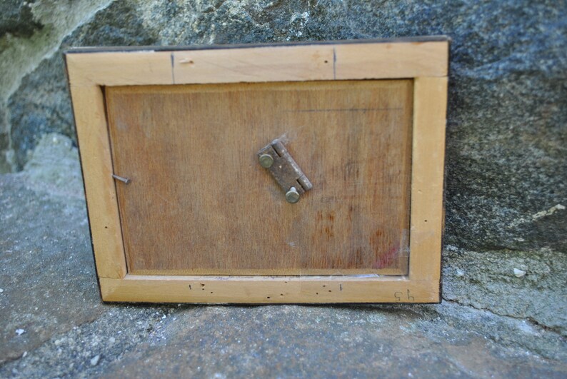 May include: The back of a wooden picture frame with a metal hinge. The frame is light brown and has a dark brown border. There are some small holes in the wood.