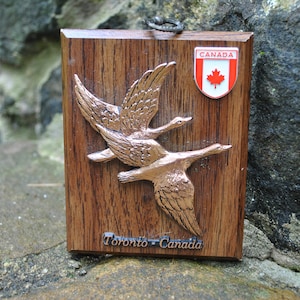 May include: A wooden plaque with a carved image of two geese in flight. The plaque is decorated with a small metal Canadian flag and the text "Toronto - Canada".
