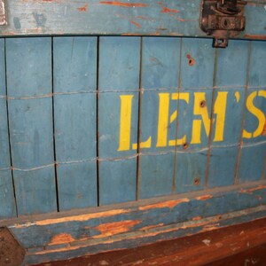May include: A blue wooden crate with a yellow painted sign that reads "LEM'S". The crate has a worn and weathered appearance, with visible signs of age and use.