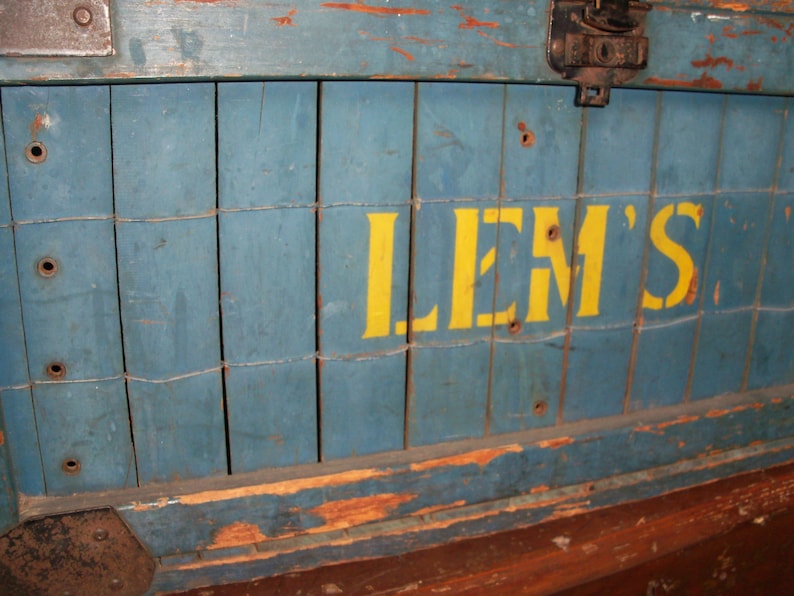 May include: A blue wooden crate with a yellow painted sign that reads "LEM'S". The crate has a worn and weathered appearance, with visible signs of age and use.
