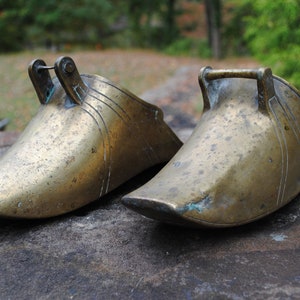 May include: Two antique brass stirrups, one with a decorative design, on a stone surface.