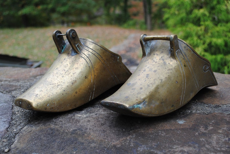 May include: Two antique brass stirrups, one with a decorative design, on a stone surface.