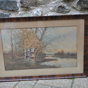 May include: A framed watercolor painting depicting a forest scene with a river in the foreground. The trees are bare and the leaves are a muted brown and yellow. The river is a light blue and reflects the sky. The frame is dark brown wood with a grain pattern.
