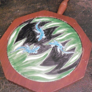 May include: A wooden octagonal cheese board with a ceramic tile insert. The tile features a green and white background with black and blue stylized lizards.