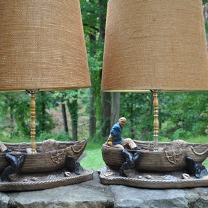 May include: A pair of vintage table lamps with a nautical theme. Each lamp features a brown, woven lampshade and a base depicting a man in a boat with a fishing net. The boats are made of a brown, textured material.