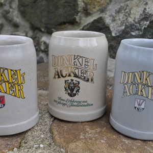 May include: Three ceramic beer steins with a white glaze and gold lettering. The steins feature the text "Dinkel Acker" and a crest with the letters "CD".