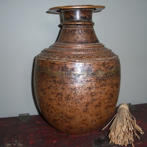 Può includere: Un grande vaso di colore rame con un corpo largo e rotondo e un collo stretto. Il vaso ha una fascia decorativa intorno al centro e un piccolo coperchio rotondo.