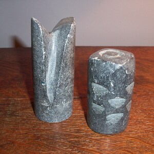 May include: Two gray stone chess pieces. One piece is a tall, slender tower with a pointed top. The other piece is a shorter, rounder cylinder with a flat top.