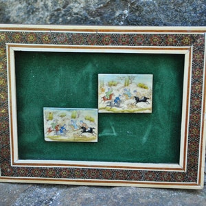 May include: A framed artwork with a green background and two small ivory tiles depicting scenes of people riding horses. The frame is made of wood and has a decorative mosaic pattern.