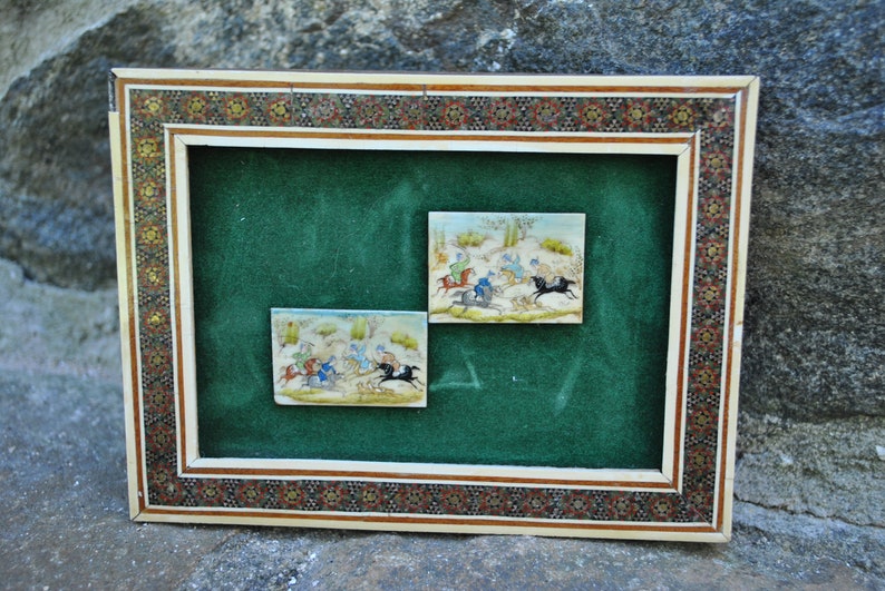 May include: A framed artwork with a green background and two small ivory tiles depicting scenes of people riding horses. The frame is made of wood and has a decorative mosaic pattern.