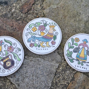 Puede incluir: Tres posavasos redondos de cerámica con diseños pintados de colores. El primer posavasos presenta un pájaro azul y amarillo con una corona, el segundo posavasos presenta una mujer que lleva dos jarras y el tercer posavasos presenta un jarrón con flores y un pájaro. Los tres posavasos tienen la palabra "SKYPOS" escrita en ellos.