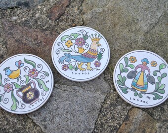Puede incluir: Tres posavasos redondos de cerámica con diseños pintados de colores. El primer posavasos presenta un pájaro azul y amarillo con una corona, el segundo posavasos presenta una mujer que lleva dos jarras y el tercer posavasos presenta un jarrón con flores y un pájaro. Los tres posavasos tienen la palabra "SKYPOS" escrita en ellos.