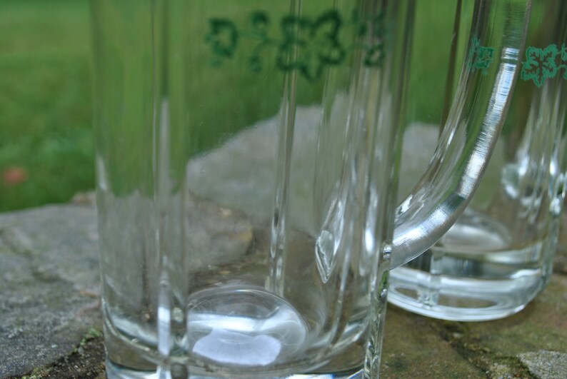 May include: Two clear glass beer mugs with handles. The mugs have a green inscription on the side.