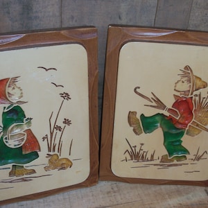 May include: Two framed wood plaques with raised relief images of children. The plaque on the left features a girl in a green dress and red and white hat holding a basket. The plaque on the right features a boy in a green and red outfit carrying a basket and a stick.