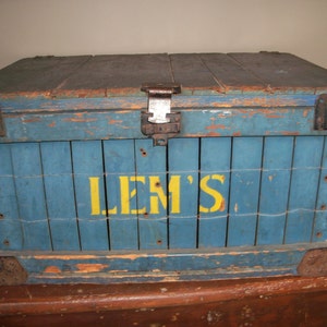 May include: A vintage wooden crate painted blue with yellow lettering that reads "LEM'S". The crate has metal hinges and clasps.