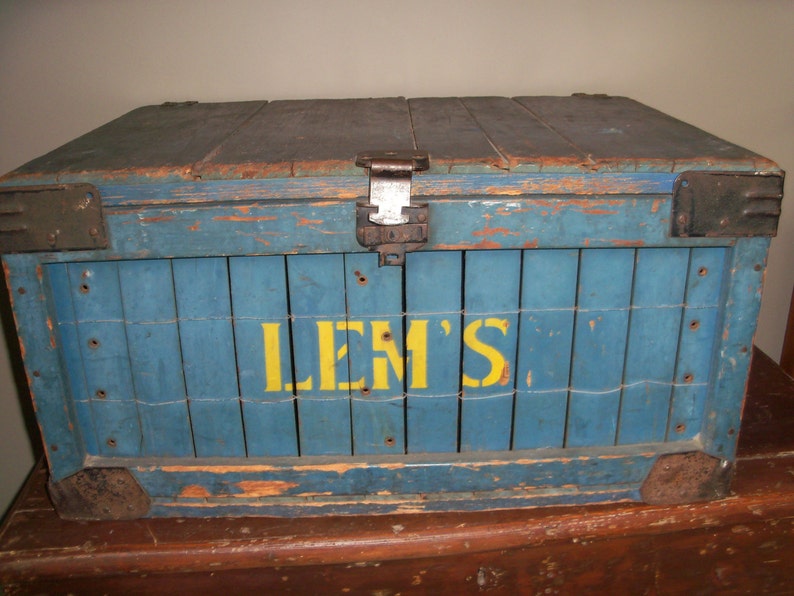May include: A vintage wooden crate painted blue with yellow lettering that reads "LEM'S". The crate has metal hinges and clasps.