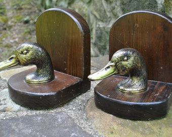 May include: A pair of wooden bookends with brass duck head finials. The bookends are dark brown with a natural wood grain. The duck heads are detailed and realistic.