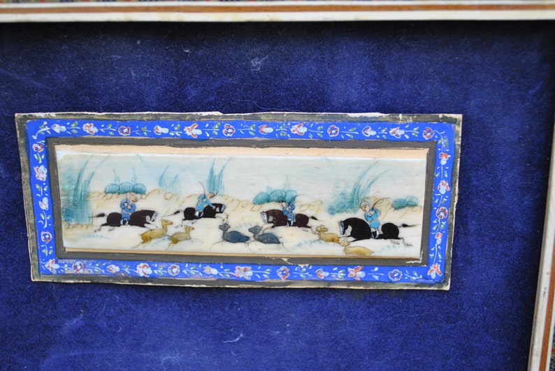 May include: A framed miniature painting on ivory depicting a hunting scene. The painting shows four figures on horseback, chasing after deer in a landscape with tall grasses. The painting is framed in a decorative border with floral motifs.