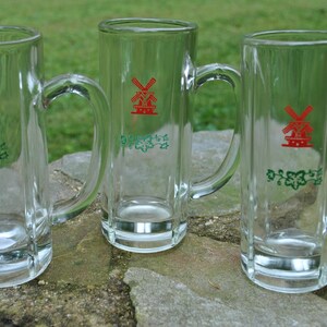 May include: Three clear glass beer mugs with handles. Each mug has a red windmill logo and green foliage design. The text "De Molen" is printed on each mug.