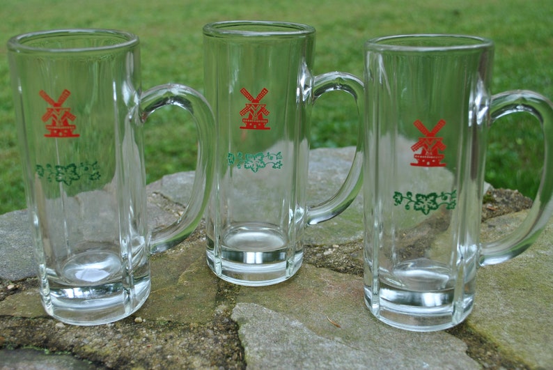 May include: Three clear glass beer mugs with handles. Each mug has a red windmill logo and green foliage design. The text "De Molen" is printed on each mug.