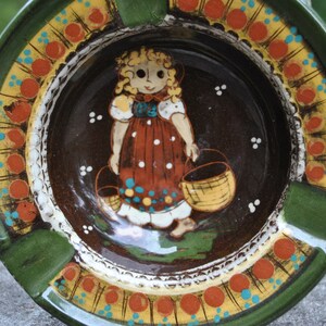 Op de afbeelding: Vintage keramische asbak met een handgeschilderd ontwerp. De asbak heeft een groene rand en een centraal beeld van een meisje in een jurk met manden. Het ontwerp bevat oranje, gele en blauwe accenten.