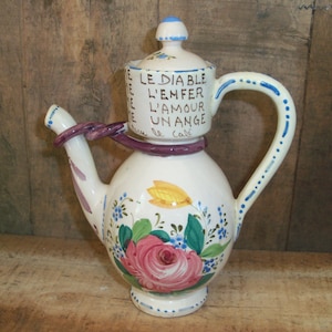 May include: White ceramic teapot with a purple handle and a lid that says "Le Diable L'Enfer L'Amour Un Ange" in French. The teapot is decorated with a pink rose, yellow flower, and blue flowers.