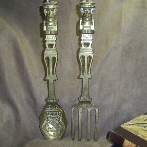 May include: A set of two gold-toned metal utensils, a spoon and a fork, with decorative carvings of a human face on the handles. The utensils are designed in a stylized, almost tribal, fashion.