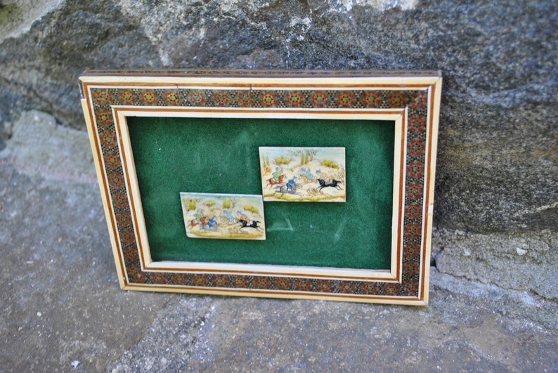 May include: Two framed miniature paintings depicting scenes of horseback riding in a desert landscape. The paintings are set against a green velvet background and framed with a decorative ivory and wood frame.