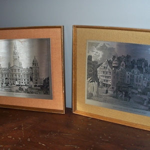 May include: Two framed black and white prints of historical cityscapes. The print on the left shows a building with a tall tower and the print on the right shows a street scene with buildings.