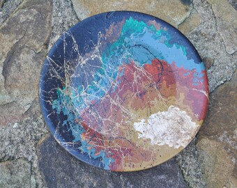 Puede incluir: Un plato redondo de cerámica con un diseño abstracto colorido. El plato está pintado en tonos de azul, verde, marrón y dorado. El diseño presenta un patrón de remolino con líneas finas y salpicaduras de pintura.