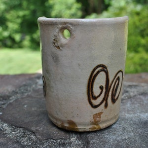K&ouml;nnte beinhalten: Eine wei&szlig;e Keramiktasse mit einem braunen Wirbelmuster. Die Tasse hat ein Loch an der Seite zum Aufh&auml;ngen.