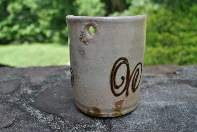 K&ouml;nnte beinhalten: Eine wei&szlig;e Keramiktasse mit einem braunen Wirbelmuster. Die Tasse hat ein Loch an der Seite zum Aufh&auml;ngen.