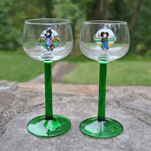 Puede incluir: Dos copas de vino de vidrio transparente con tallos y bases verdes. Cada copa tiene una pegatina colorida con la representación de una persona con ropa tradicional.
