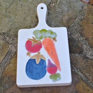 May include: White ceramic cutting board with a painted design of vegetables. The vegetables include a red tomato, a blue eggplant, a purple beet, and an orange carrot.