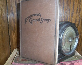 May include: A brown book with the title "Rodeheaver's Gospel Songs" printed on the cover. The book is sitting on a wooden shelf next to a vintage record player.