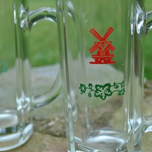May include: Two clear glass beer mugs with handles. Each mug has a green vine design and a red windmill graphic with the text "De Molen".
