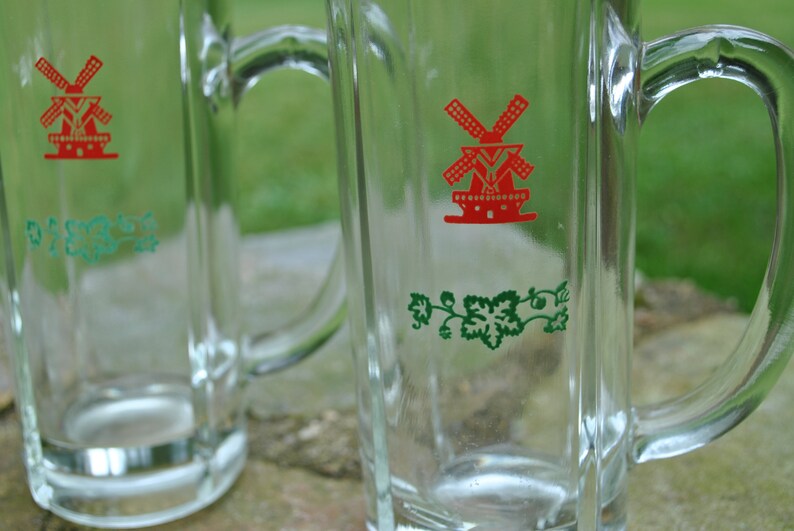 May include: Two clear glass beer mugs with handles. Each mug has a green vine design and a red windmill graphic with the text "De Molen".