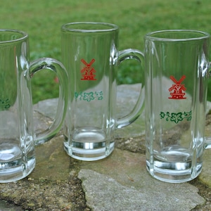 May include: Three clear glass beer mugs with a red windmill and green vines design. The mugs have a handle on the right side.