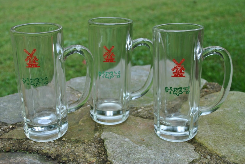 May include: Three clear glass beer mugs with a red windmill and green vines design. The mugs have a handle on the right side.