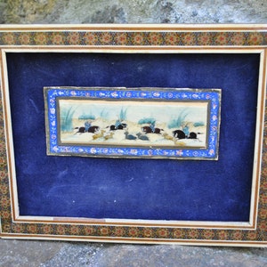 May include: A framed miniature painting depicting a hunting scene with figures on horseback pursuing animals. The painting is set against a light blue background and framed in a decorative wooden frame with a colorful mosaic border.