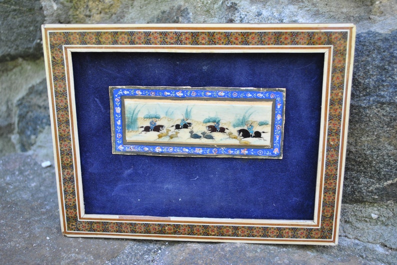 May include: A framed miniature painting depicting a hunting scene with figures on horseback pursuing animals. The painting is set against a light blue background and framed in a decorative wooden frame with a colorful mosaic border.