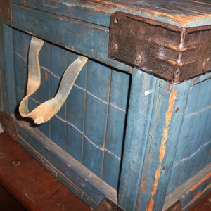May include: A vintage wooden crate with a blue paint finish and a metal latch. The crate has a leather strap handle and is showing signs of wear and tear.