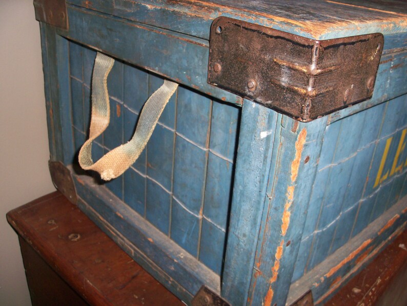 May include: A vintage wooden crate with a blue paint finish and a metal latch. The crate has a leather strap handle and is showing signs of wear and tear.