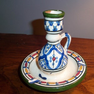 Puede incluir: Un candelero de cerámica blanca con un patrón geométrico azul y verde. El candelero tiene una pequeña asa y se asienta sobre un platillo a juego.