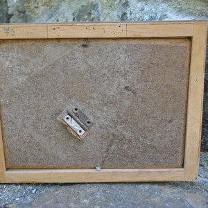 May include: The back of a wooden picture frame with a number "48" written on the top edge. The frame has a brown paper backing and a metal hinge attached to the back.