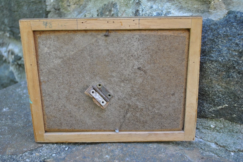 May include: The back of a wooden picture frame with a number "48" written on the top edge. The frame has a brown paper backing and a metal hinge attached to the back.